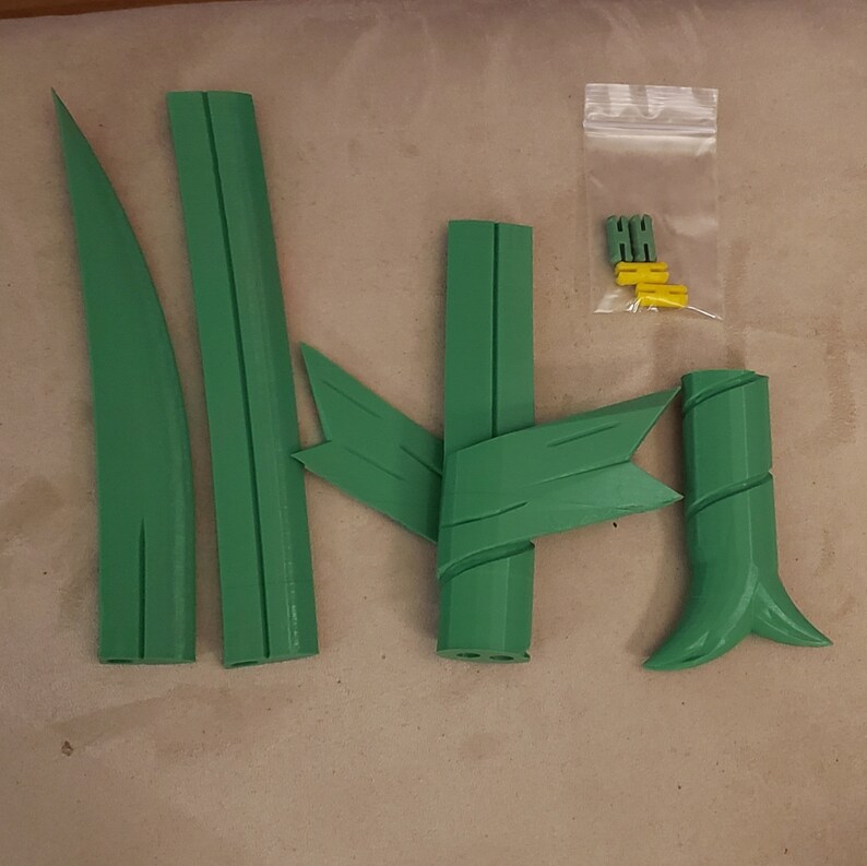 K&ouml;nnte beinhalten: Gr&uuml;ne 3D-gedruckte Komponenten einer Pflanze, einschlie&szlig;lich Bl&auml;tter und Stiele, sind auf einer neutralen Oberfl&auml;che angeordnet. Ein kleiner Beutel enth&auml;lt zus&auml;tzliche gelbe und gr&uuml;ne Teile. Die Teile scheinen f&uuml;r ein Dekorations- oder Bastelprojekt bestimmt zu sein.