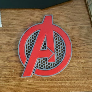 May include: A red and gray plastic Avengers logo with a honeycomb pattern. The logo is a circle with the letter A in the center.