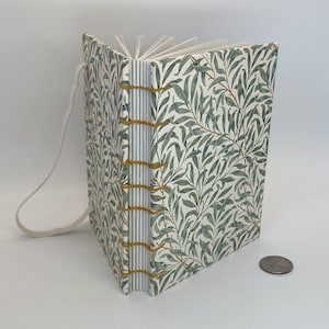 Op de afbeelding: Een handgemaakt notitieboek met een botanische print op de kaft met groene bladeren en takken op een witte achtergrond. De pagina's zijn gebonden met gouden draad en een crèmekleurige lintsluiting. Het notitieboek is open en toont de lege pagina's.