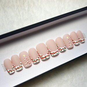 May include: A set of ten press-on nails with a matte nude base. The tips feature a white French manicure style with a red ribbon and green leaf pattern. Some nails include a small, stylized white Christmas tree design with a yellow star.