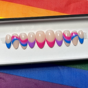 May include: A set of press-on nails with a nude base and colorful tips. The tips feature designs in blue, pink, and purple, arranged in a pattern. The nails are oval-shaped and presented in a white box, with a rainbow flag in the background.