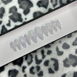 May include: A set of ten white press-on nails with iridescent rhinestones.