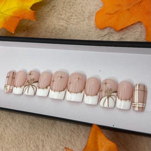 May include: A set of ten press-on nails featuring a nude base with white French tips. Some nails include gold line art of pumpkins and starburst accents, while others feature a brown and white plaid pattern on the sides of the nail tips.