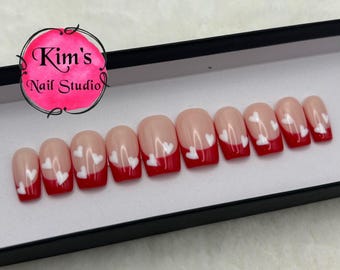 May include: A set of press-on nails with a red and white heart design. The nails have a nude base with a red French tip and white heart accents. The image includes the text "Kim's Nail Studio".