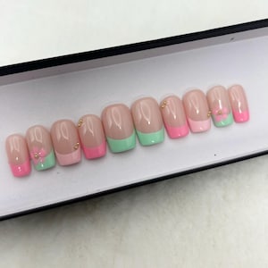 May include: A set of artificial nails with a French manicure design. The nails feature a clear base, pink and mint green tips, and small gold accents. Some nails have a small flower design.