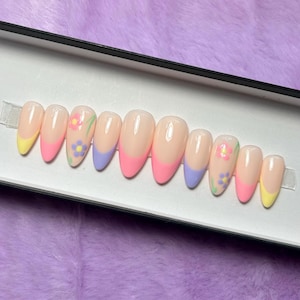 May include: A set of artificial nails in a box. The nails have a light pink base with colorful tips and floral designs. The tips are painted in yellow, pink, and blue. Some nails feature small flower decals.