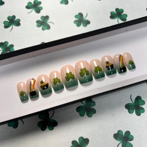 May include: A set of press-on nails with a St. Patrick's Day theme. Each nail features a gradient green base, with designs including shamrocks, a pot of gold, and a rainbow. The nails are arranged on a white surface with green shamrock patterns.