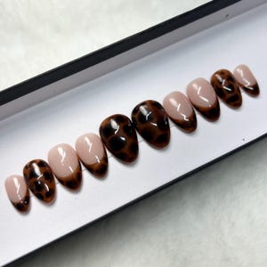 May include: A set of ten almond-shaped press-on nails featuring a glossy finish. Each nail has a nude base color with a dark brown and black leopard print pattern applied as a French tip design, arranged in a row inside a white display box.