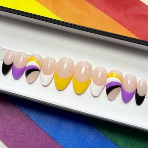 May include: A set of oval-shaped press-on nails with a clear base and various colored tips. The tips are painted in black, white, yellow, and purple, with some nails featuring non-binary flag designs. The nails are displayed in a white box against a rainbow background.