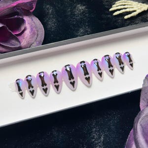May include: A set of oval-shaped, iridescent press-on nails. Each nail features a black bone design and a black heart at the tip. The nails are arranged in a row, showcasing a gothic or Halloween theme.