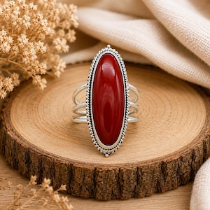 May include: A silver ring featuring a large, oval-shaped, smooth red stone set in a decorative twisted rope metal border. The ring has a multi-band shank design that splits into three separate silver wires on each side of the central stone setting.