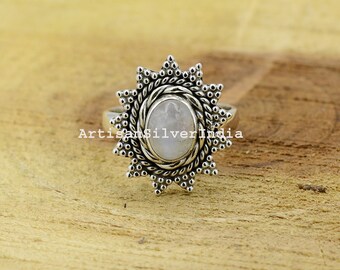 May include: A silver ring featuring an oval, pale gemstone at its centre. The stone is framed by a detailed, sunburst design. The ring is set against a wooden surface, with the text "ArtisanSilverIndia" visible.