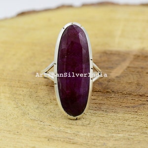 Pode incluir: Um anel de prata com uma grande pedra preciosa ovalada de cor púrpura. O anel tem um design simples com uma banda fina e uma configuração de bezel para a pedra preciosa.