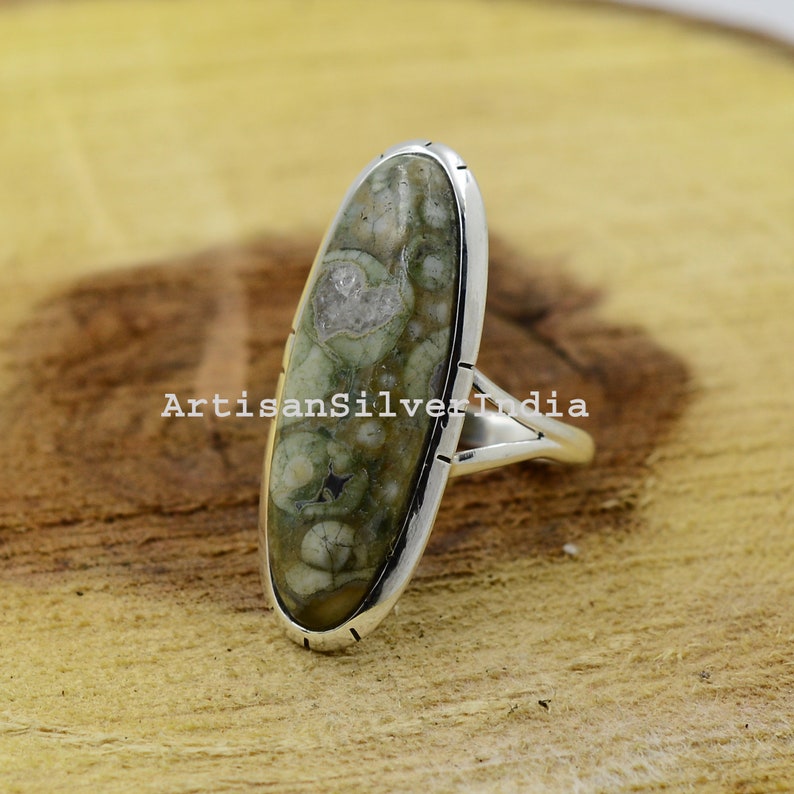 K&ouml;nnte beinhalten: Ein silberner Ring mit einem ovalen, gr&uuml;n-wei&szlig;en Stein, der in der Mitte eingesetzt ist. Der Ring hat ein schlichtes Band und liegt auf einer Holzoberfl&auml;che.