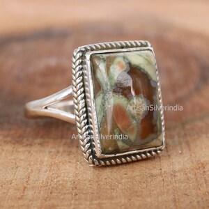 May include: A silver ring featuring a rectangular, green, brown and white stone set within a rope-like border.