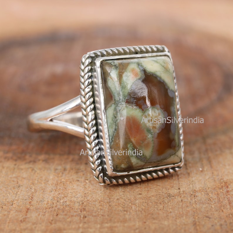 May include: A silver ring featuring a rectangular, green, brown and white stone set within a rope-like border.