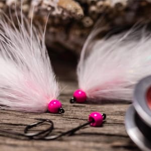 Könnte beinhalten: Vier weiße und rosafarbene Fliegenfischerköder mit rosafarbenen Perlen an Haken. Die Köder liegen auf einer Holzoberfläche neben einer Spule mit Angelschnur.
