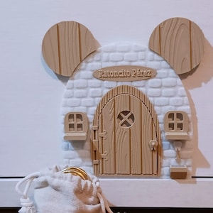 May include: A whimsical wooden mouse door with a brick facade, two windows, and a door. The door has the text "Ratoncito Pérez" above it. A small white drawstring bag filled with gold coins sits in front of the door.