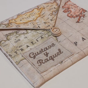 Op de afbeelding: Een beige stoffen envelop met een wereldkaartprint. De envelop heeft een bruine knoopsluiting en een gepersonaliseerd geborduurd label met de tekst "Gustavo y Raquel".