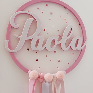 Può includere: Cerchio di legno rosa con il nome "Paola" scritto in lettere bianche. Il cerchio è decorato con stelle glitter rosa e ha nastri rosa e bianchi che pendono dal fondo.