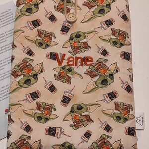 Puede incluir: Una bolsa de tela beige con cierre de cordón. La bolsa está decorada con un patrón repetido de personajes de dibujos animados verdes que sostienen tazas de café. El nombre "Vane" está bordado en hilo naranja en la parte delantera de la bolsa.