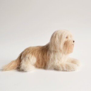 May include: A plush, cream-colored dog figurine with light brown markings, lying down on a white surface. The dog has long, shaggy fur and is posed in a relaxed position. The figurine is a decorative item.