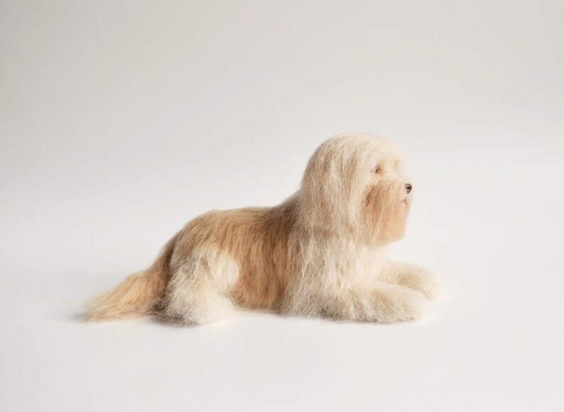 May include: A plush, cream-colored dog figurine with light brown markings, lying down on a white surface. The dog has long, shaggy fur and is posed in a relaxed position. The figurine is a decorative item.