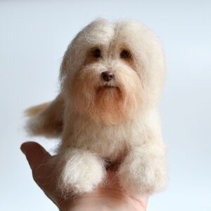 May include: A handmade, light beige dog figurine with a fluffy, textured appearance. The dog has a brown nose and eyes, and is resting on a hand. The figurine is made of wool or a similar material.