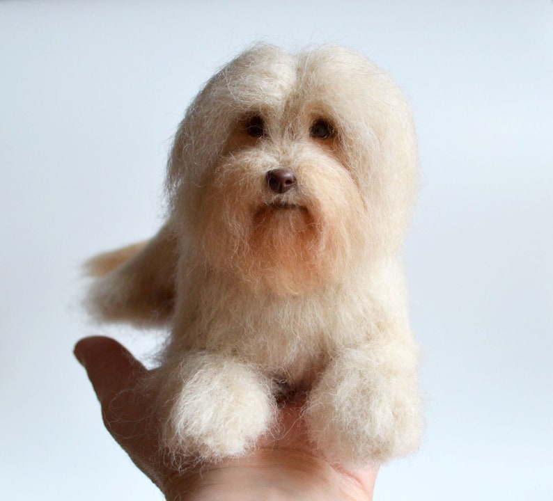 May include: A handmade, light beige dog figurine with a fluffy, textured appearance. The dog has a brown nose and eyes, and is resting on a hand. The figurine is made of wool or a similar material.