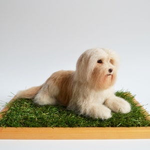 May include: A handmade, light brown and white dog figurine resting on a bed of green faux grass within a wooden frame. The dog has long, fluffy fur and is lying down, appearing relaxed. The background is plain white.