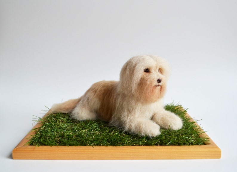 May include: A handmade, light brown and white dog figurine resting on a bed of green faux grass within a wooden frame. The dog has long, fluffy fur and is lying down, appearing relaxed. The background is plain white.