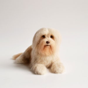 May include: A handcrafted, light beige dog figurine, lying down with its front paws extended. The dog has long, fluffy fur, a brown nose, and a peach-colored beard. The background is plain white.