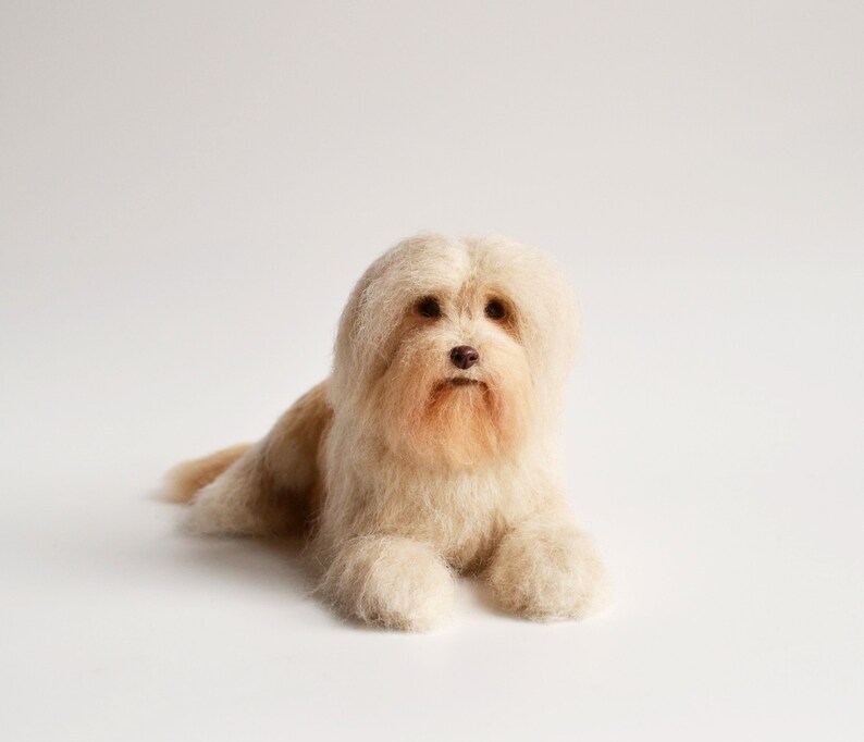 May include: A handcrafted, light beige dog figurine, lying down with its front paws extended. The dog has long, fluffy fur, a brown nose, and a peach-colored beard. The background is plain white.