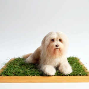 May include: A handmade, light beige dog figurine with long, fluffy fur, lying on a bed of green artificial grass. The dog rests on a wooden tray, creating a natural and charming display. The background is plain white.