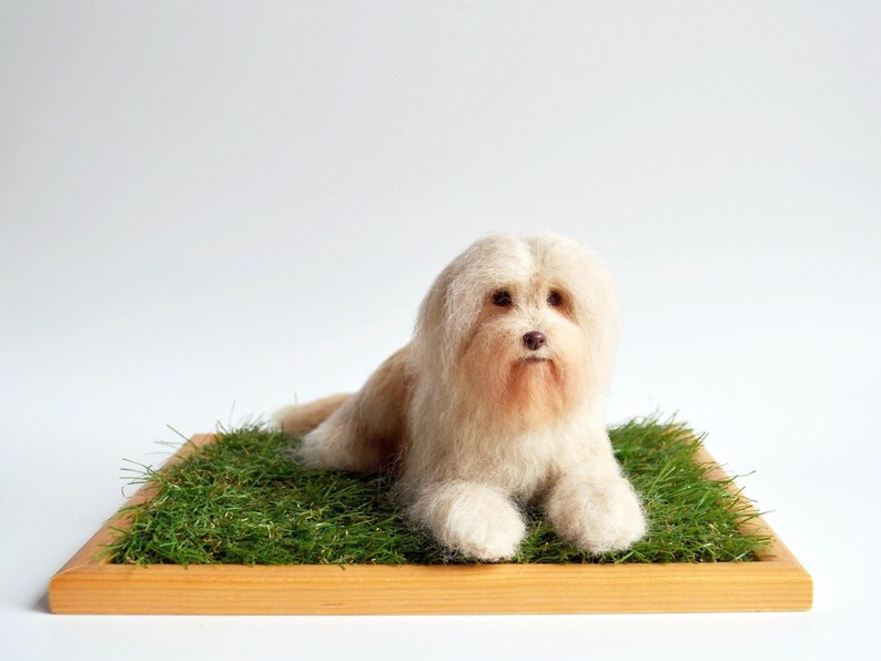 May include: A handmade, light beige dog figurine with long, fluffy fur, lying on a bed of green artificial grass. The dog rests on a wooden tray, creating a natural and charming display. The background is plain white.