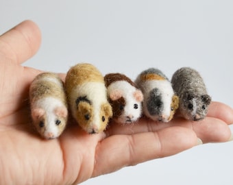 May include: Five miniature guinea pig figurines in various colors, including tan, brown, and gray, are displayed on a person's open palm. Each figurine has small, dark eyes and is made of a soft, textured material.