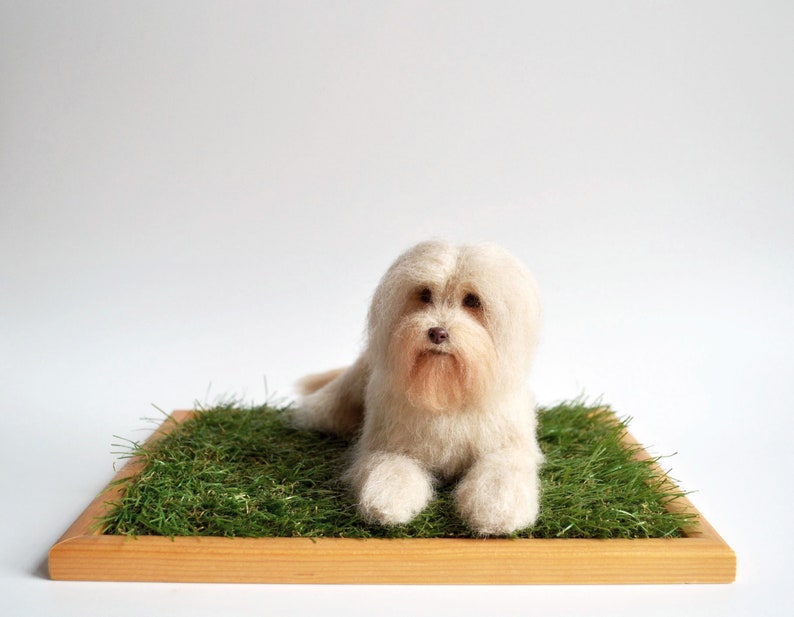 May include: A handmade, off-white dog figurine with a fluffy coat, lying on a bed of green artificial grass. The dog is set in a wooden frame, creating a miniature scene. The dog's features are detailed, with a small, brown nose and expressive eyes.
