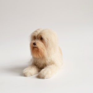 May include: A handmade, light beige dog figurine with long, flowing fur. The dog is lying down, with its front paws extended. The fur around the face and muzzle is a slightly darker shade of beige, creating a natural look. The dog has a small, brown nose.