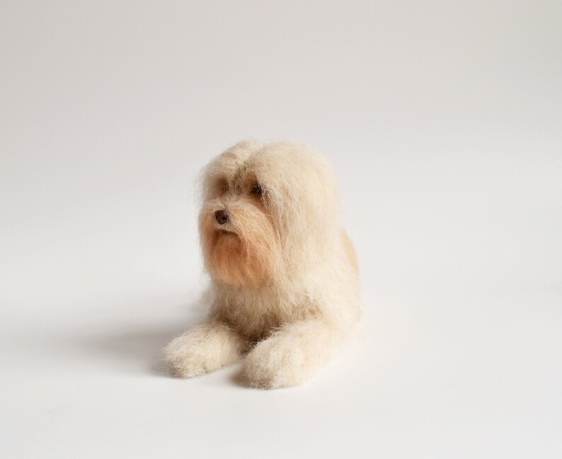 May include: A handmade, light beige dog figurine with long, flowing fur. The dog is lying down, with its front paws extended. The fur around the face and muzzle is a slightly darker shade of beige, creating a natural look. The dog has a small, brown nose.