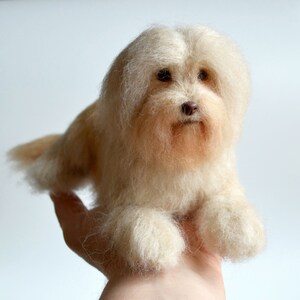 May include: A handmade, cream-colored dog figurine with a fluffy, textured appearance. The dog has a light brown nose and dark eyes, and is lying down. The figurine is held in a person's hand.