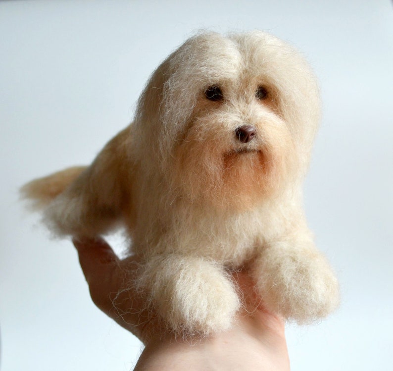 May include: A handmade, cream-colored dog figurine with a fluffy, textured appearance. The dog has a light brown nose and dark eyes, and is lying down. The figurine is held in a person's hand.