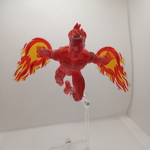 Puede incluir: Una figura de acción roja y amarilla de un superhéroe con alas de fuego. La figura está en posición de vuelo con los brazos extendidos.