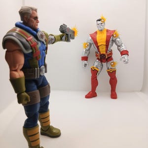 May include: Two action figures, one in blue and silver armor aiming a gun at a second figure in red, yellow, and silver armor. The figures have yellow bursts indicating impact.