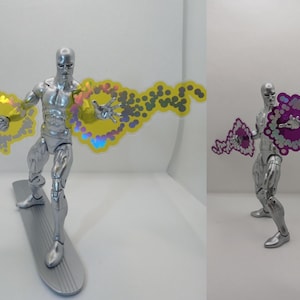 May include: A silver action figure of a character on a silver surfboard, with yellow and purple energy effects emanating from the figure's hands. The figure is posed dynamically, suggesting motion and power. The figure is a collectible item.