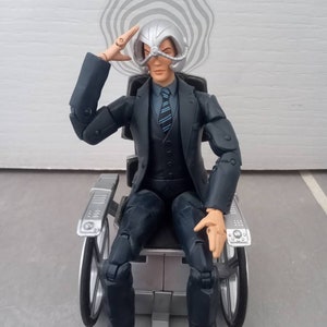 May include: A silver and black action figure of a man in a suit sitting in a wheelchair. The figure has a silver helmet with a circular design on top of his head. The figure is wearing a dark blue suit with a light blue shirt and a dark blue tie. The wheelchair is silver and black with two wheels on each side.
