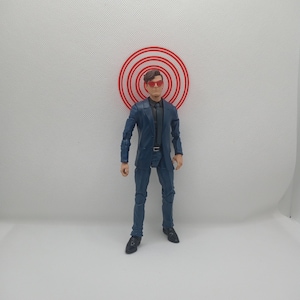 Könnte beinhalten: Eine blaue Actionfigur in Anzug und Sonnenbrille steht vor einem roten Ziel. Die Figur trägt einen dunkelblauen Anzug, schwarze Schuhe und eine rote Sonnenbrille. Das Ziel ist eine Reihe roter konzentrischer Kreise.