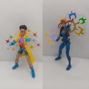 以下が含まれることがあります： 黄色いコートと青いスーツを着た2つのアクションフィギュア。それぞれ透明なプラスチック効果があります。左側のフィギュアはピンクのメガネと青い手袋をしています。右側のフィギュアは茶色のジャケットと赤いヘアバンドをしています。