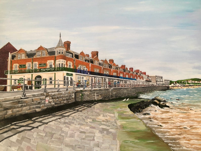 May include: A painting of a row of brick buildings along a waterfront with a stone walkway and a sandy beach in the background.