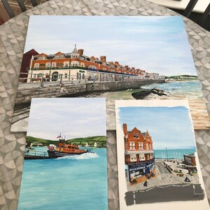 May include: Three paintings of coastal scenes. The first painting shows a row of buildings along a waterfront with a blue sky and white clouds. The second painting shows a lifeboat on the water with a blue sky and white clouds. The third painting shows a building with a sign that reads "The Fish Place" with a blue sky and white clouds.