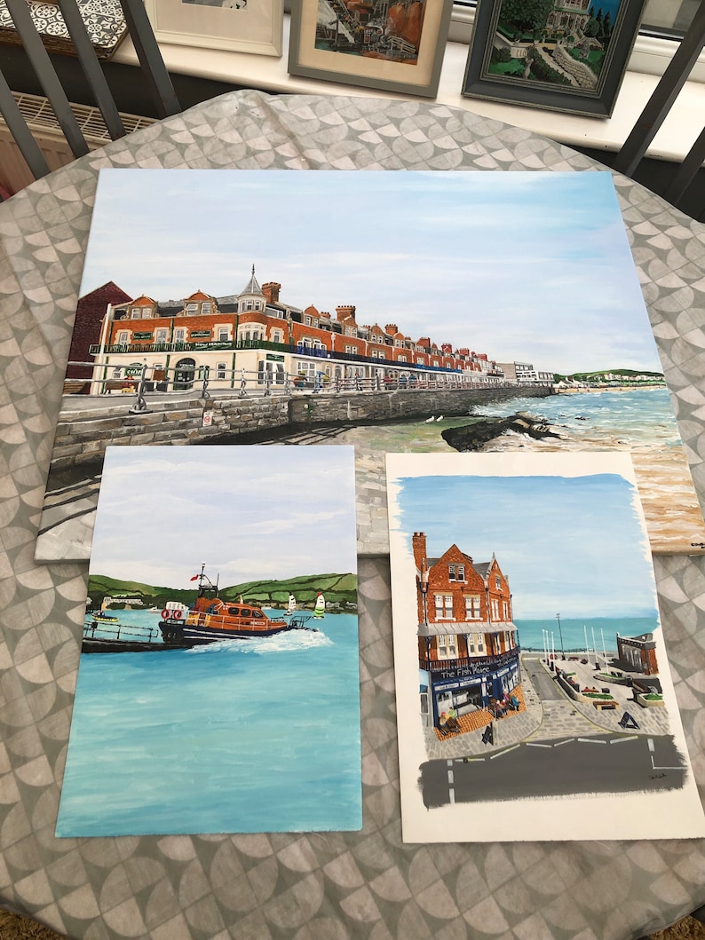 May include: Three paintings of coastal scenes. The first painting shows a row of buildings along a waterfront with a blue sky and white clouds. The second painting shows a lifeboat on the water with a blue sky and white clouds. The third painting shows a building with a sign that reads "The Fish Place" with a blue sky and white clouds.